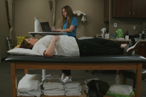 Woman lying on medical table with legs crossed, specialist in background reviewing information on a computer.